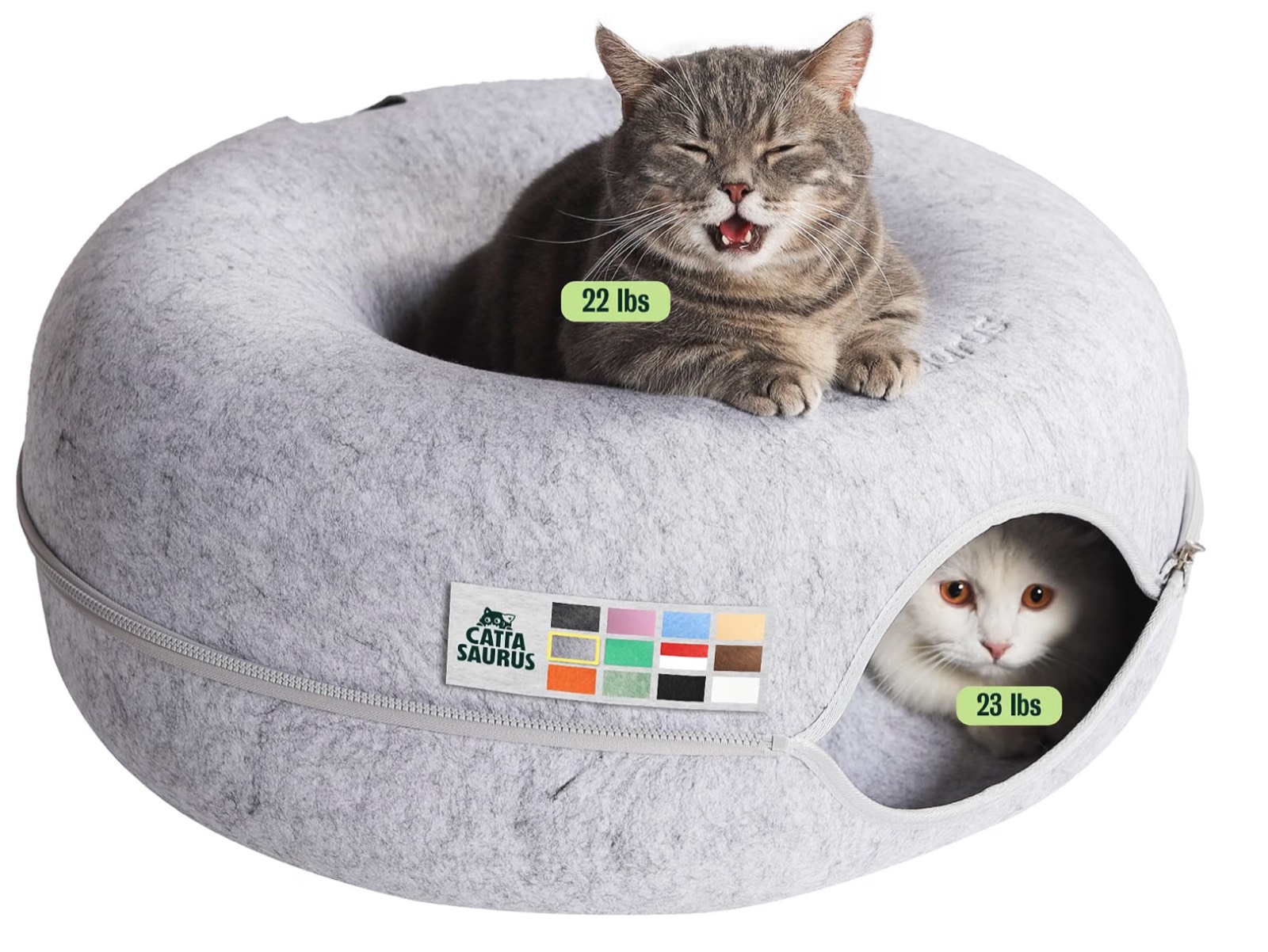 A grey felt donut cat cave with a tabby resting on top and a white cat peeking out of the tunnel.